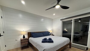 Une chambre à coucher comprenant un lit, deux lampes de chevet, un ventilateur de plafond et une porte-fenêtre coulissante donnant sur une autre pièce.