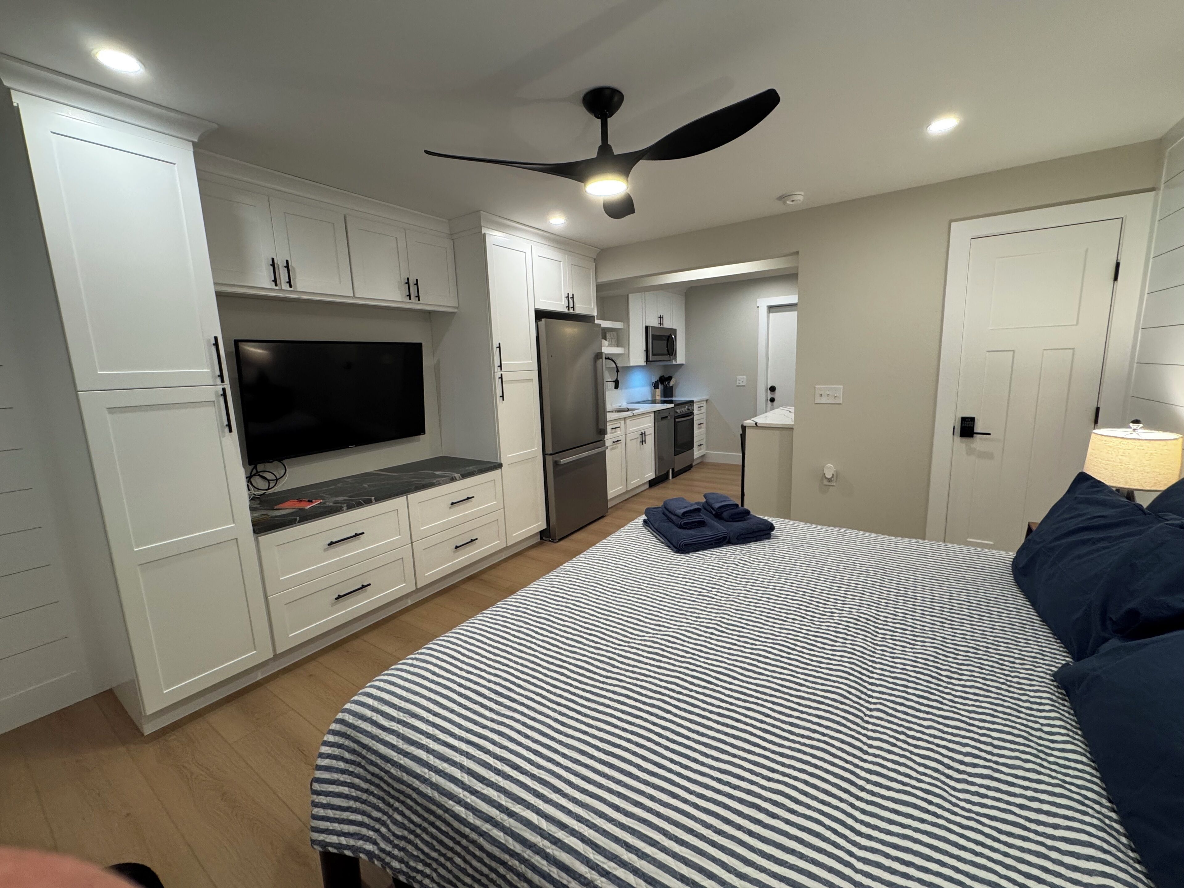 A modern bedroom with a bed, a TV, a ceiling fan, and a kitchenette in the background.