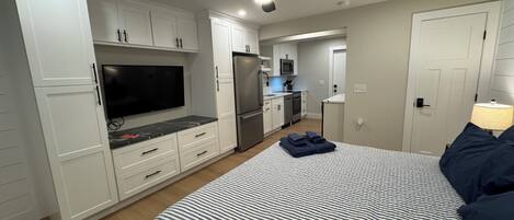 A modern bedroom with a bed, a TV, a ceiling fan, and a kitchenette in the background.