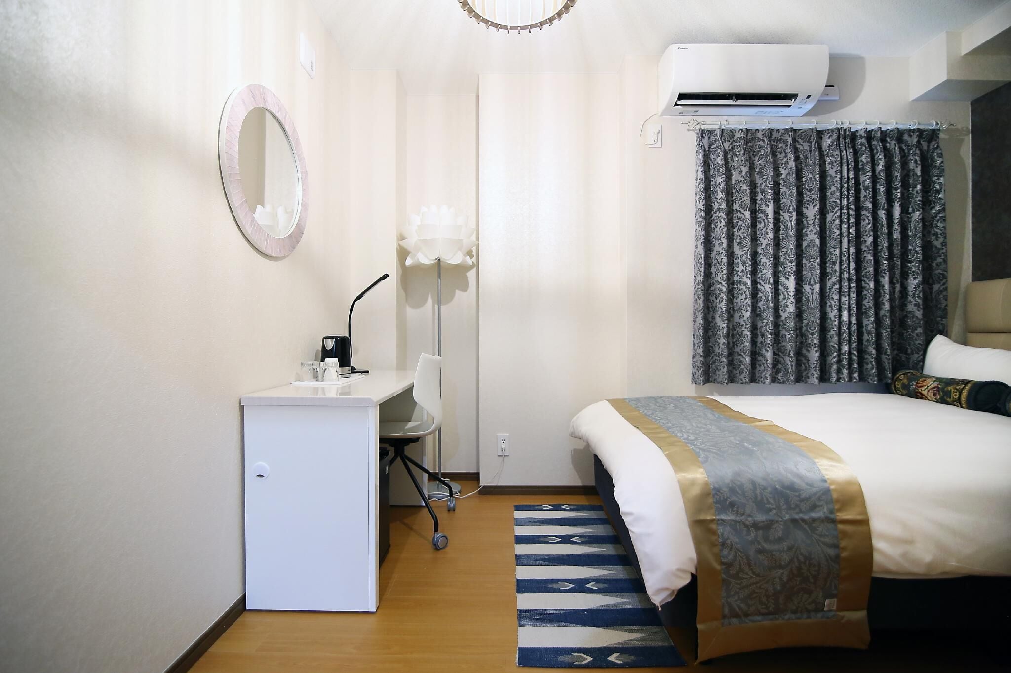 A hotel room with a bed, a desk with a chair, a small refrigerator, a mirror, and an air conditioning unit.