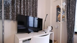 Quarto de hotel com televisão de ecrã plano sobre uma secretária branca, uma máquina de café e um cabido com um roupeiro.