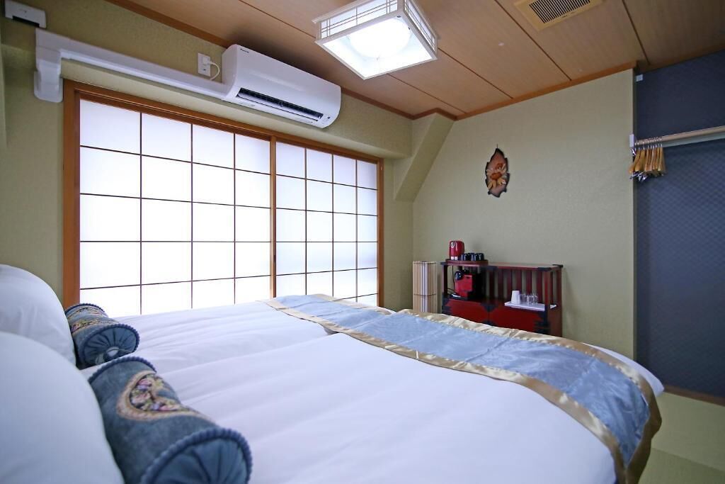 Et hotelværelse med en stor seng, en aircondition, et vindue med et gittermønster og et lille skrivebord med en rød stol.