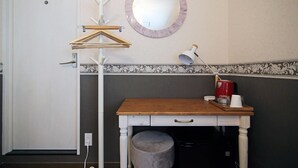Quarto pequeno com escrivaninha de madeira, espelho redondo, secador de cabelo e cafeteira.