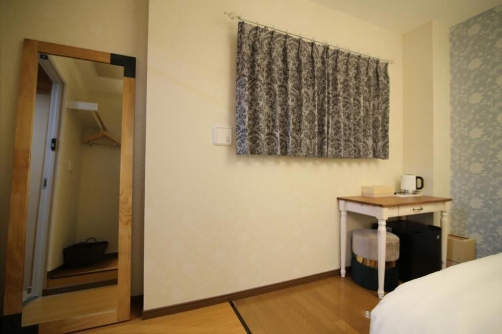 A hotel room with a wooden floor, a bed, a desk with a chair, a wardrobe, and a curtain.