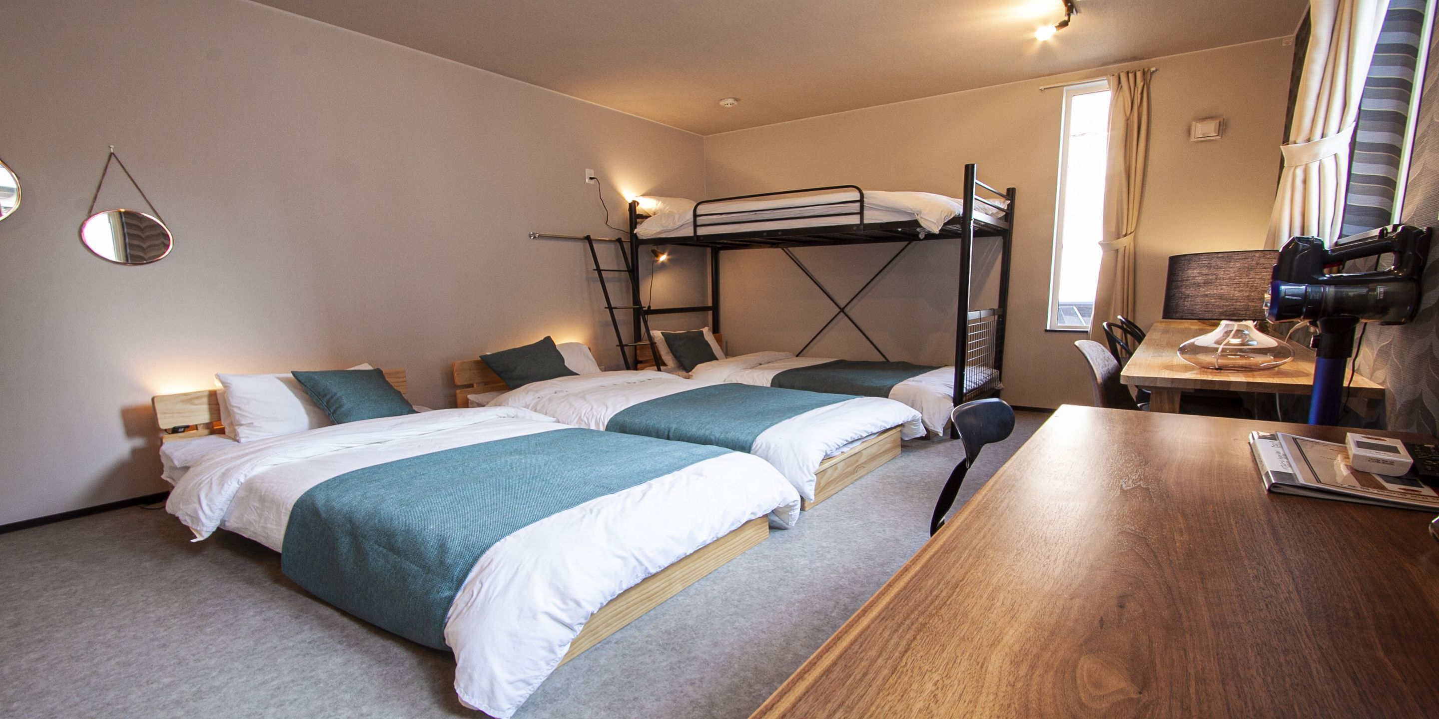 Quarto de hotel com duas camas de solteiro, uma cama beliche, uma escrivaninha de madeira e um espelho na parede.