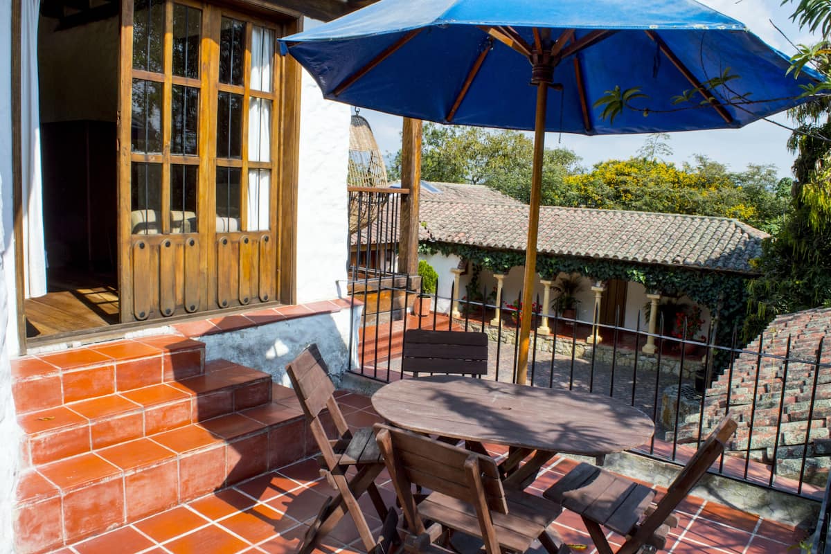 A patio with a table and chairs, a blue umbrella, and a view of a house with a tiled roof and a balcony.