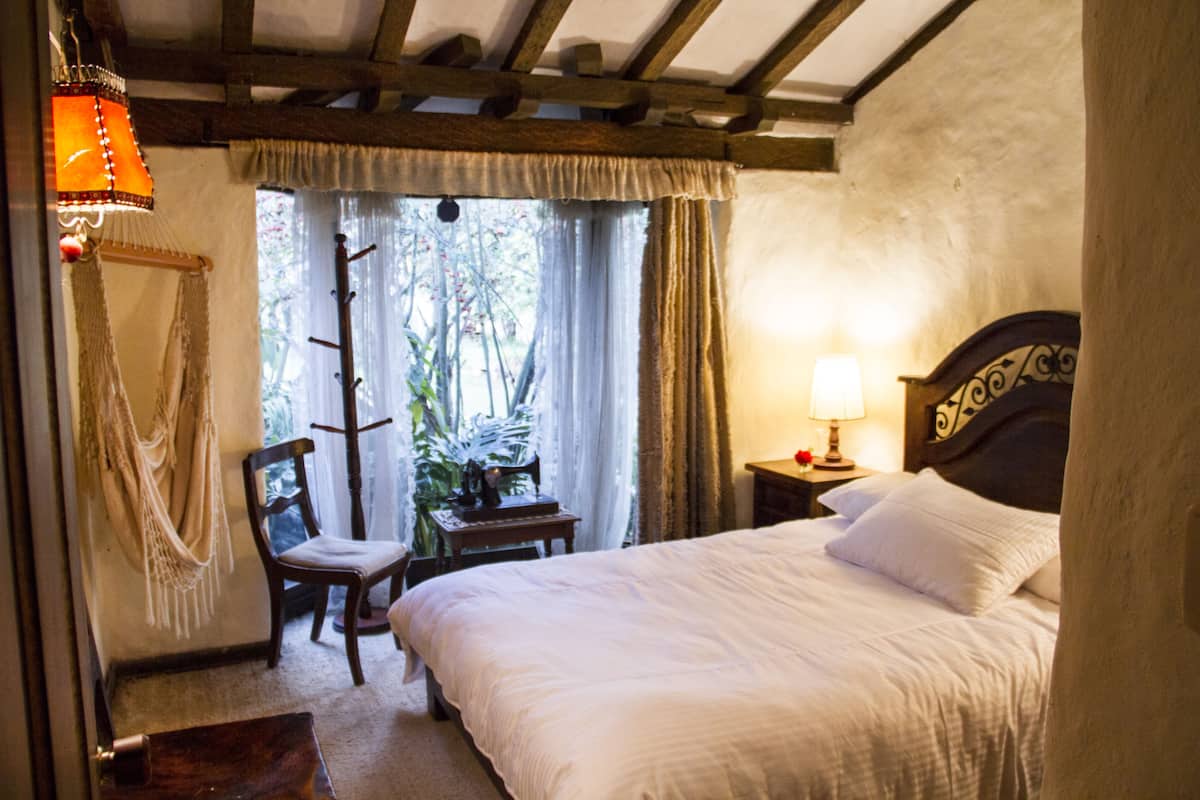 A bedroom with a bed, a chair, a nightstand, a lamp, and a window with curtains.