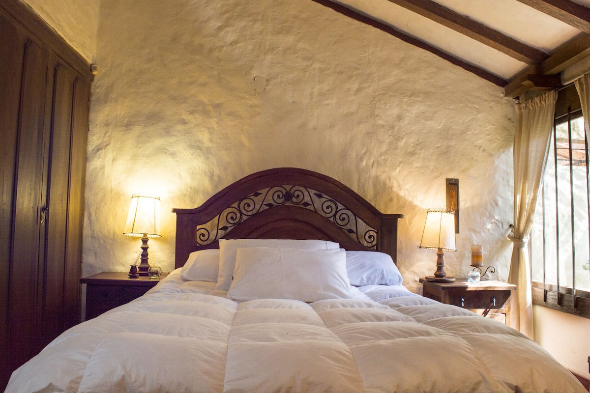 A bedroom with a large bed, two bedside tables with lamps, a wooden headboard, and a window with curtains.