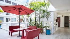 A red bench and table under a red umbrella in a courtyard with potted plants and a white building in the background. - Hotel Rimon (Cozumel)