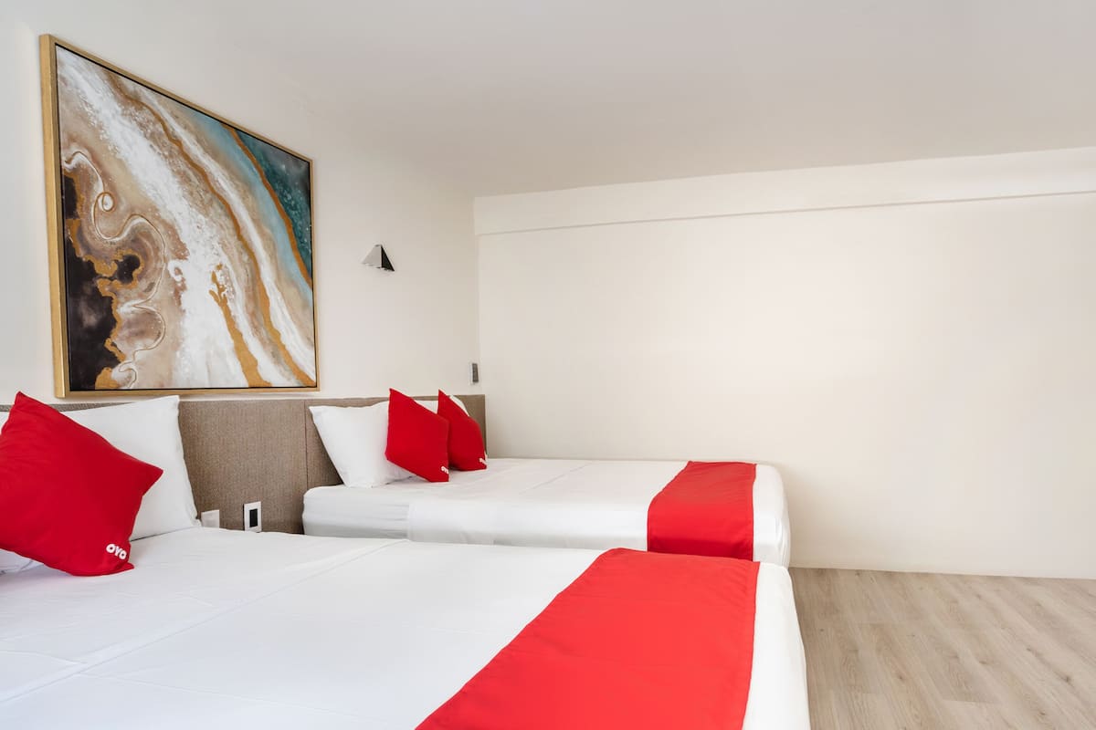 A hotel room with two beds, each with white bedding and red accents, a large abstract painting on the wall, and a wooden floor.