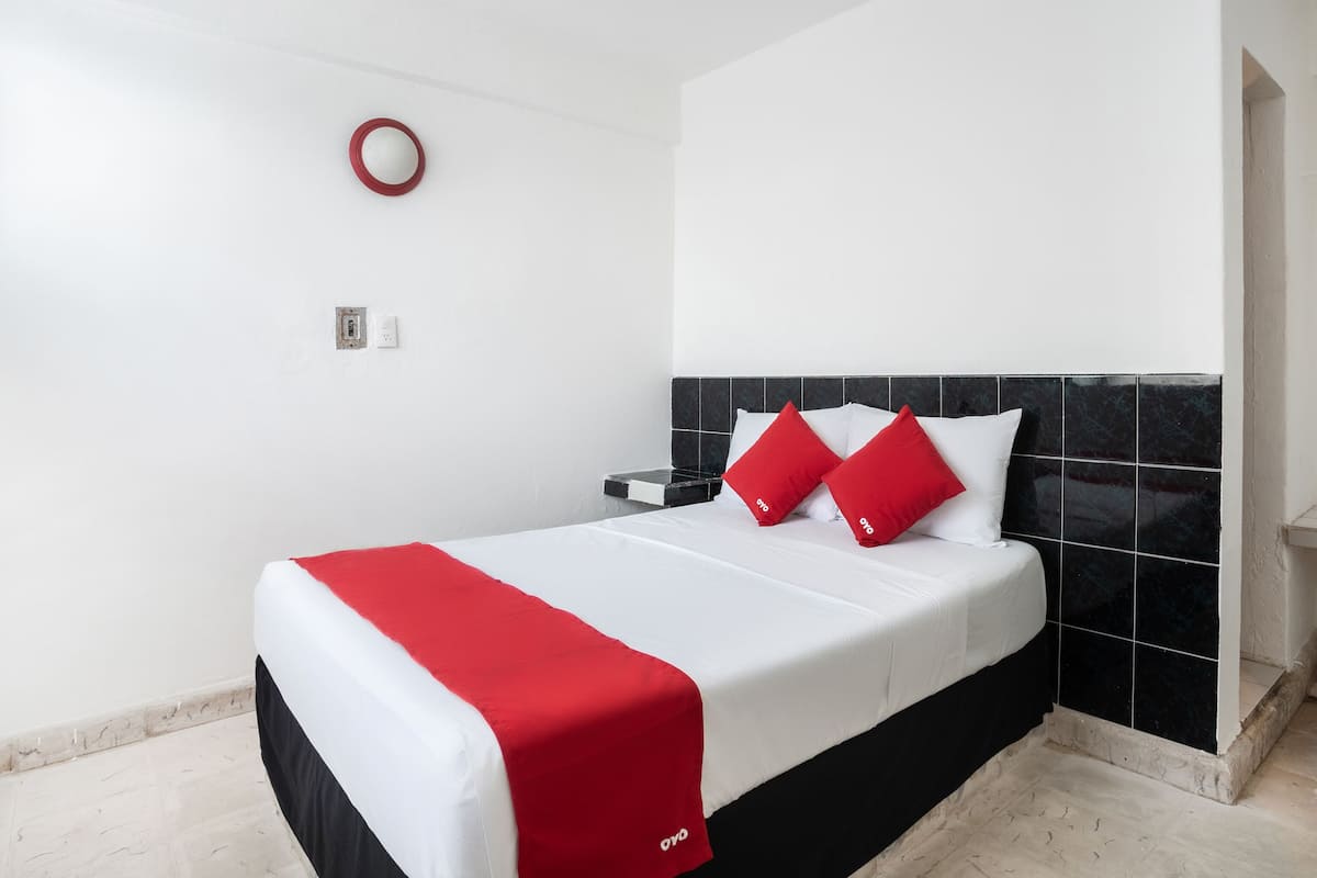 A hotel room with a bed, white walls, a black tiled headboard, and a red and white bedspread.