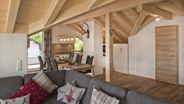 A spacious living area with a high wooden ceiling, a grey sofa with patterned cushions, a dining table with chairs, and a kitchen area visible through an open doorway.