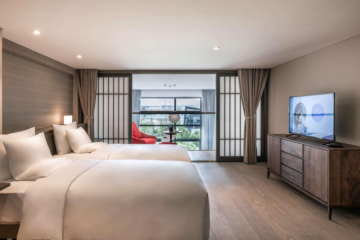 Ein Hotelzimmer mit einem großen Bett, einem Fernseher, einem Schreibtisch und Blick auf die Stadt durch große Fenster.