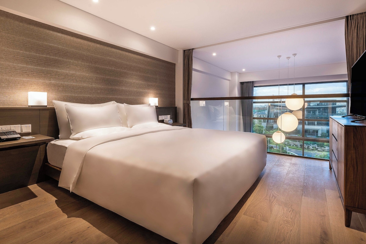 Ein modernes Hotelzimmer mit einem großen Bett, Nachttischlampen, einem Beistelltisch und Blick auf die Stadt durch große Fenster.