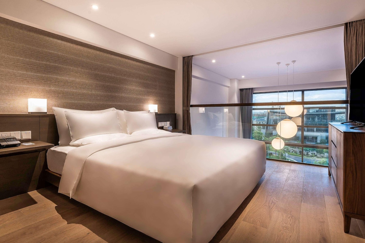 Habitación de hotel moderna con una cama grande, mesitas de noche, un escritorio y vistas a la ciudad a través de amplios ventanales.