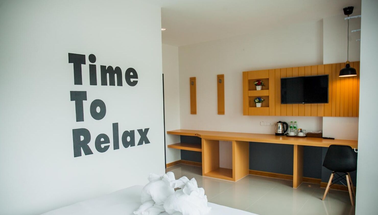 A modern room with a large white wall displaying "Time To Relax" in bold letters. There is a wooden desk with a flat-screen TV, a coffee maker, and a small shelf with decorative items. A black chair is positioned in front of the desk.