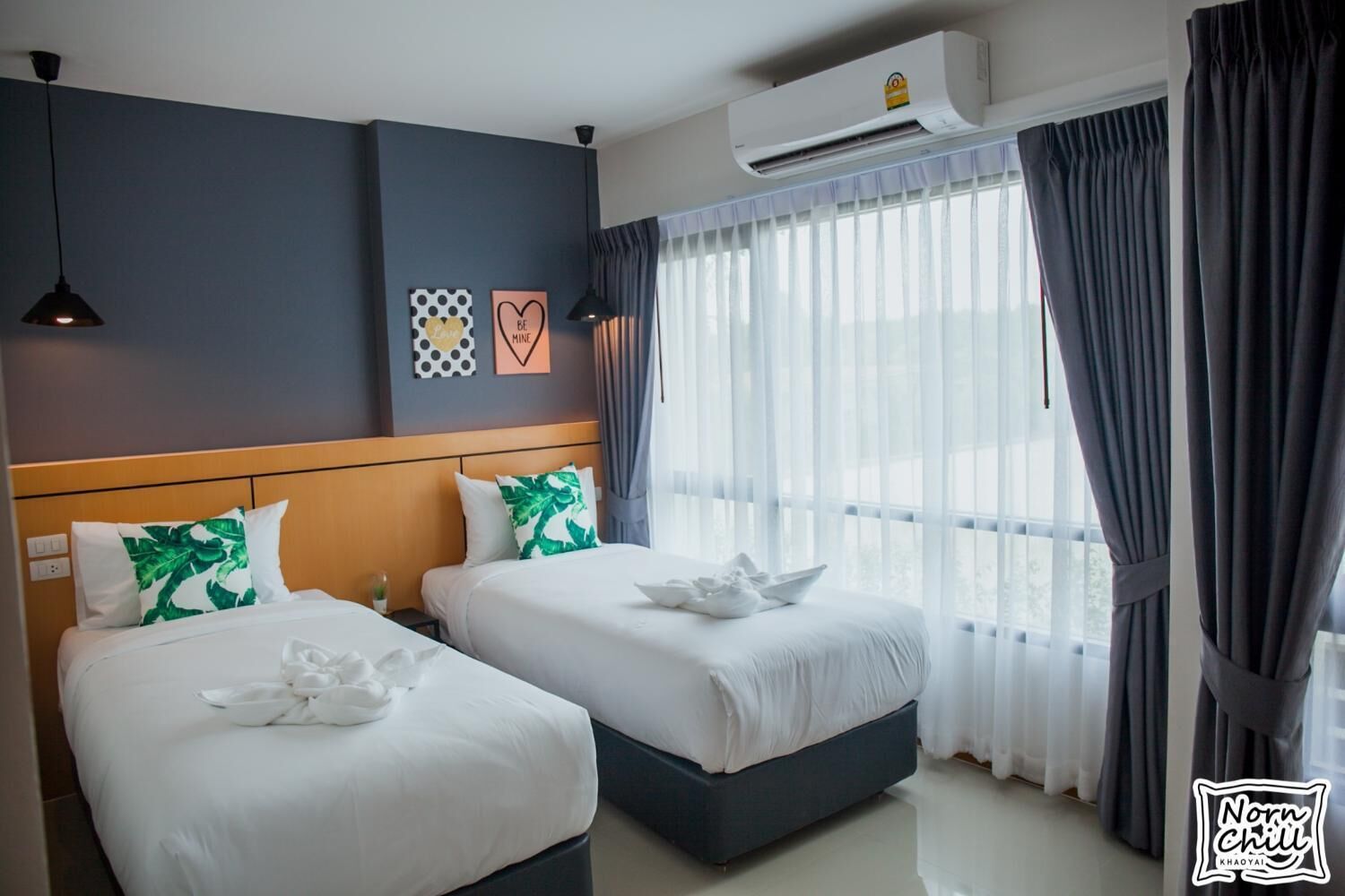 A hotel room with two beds, a wooden headboard, a large window with curtains, and an air conditioning unit.