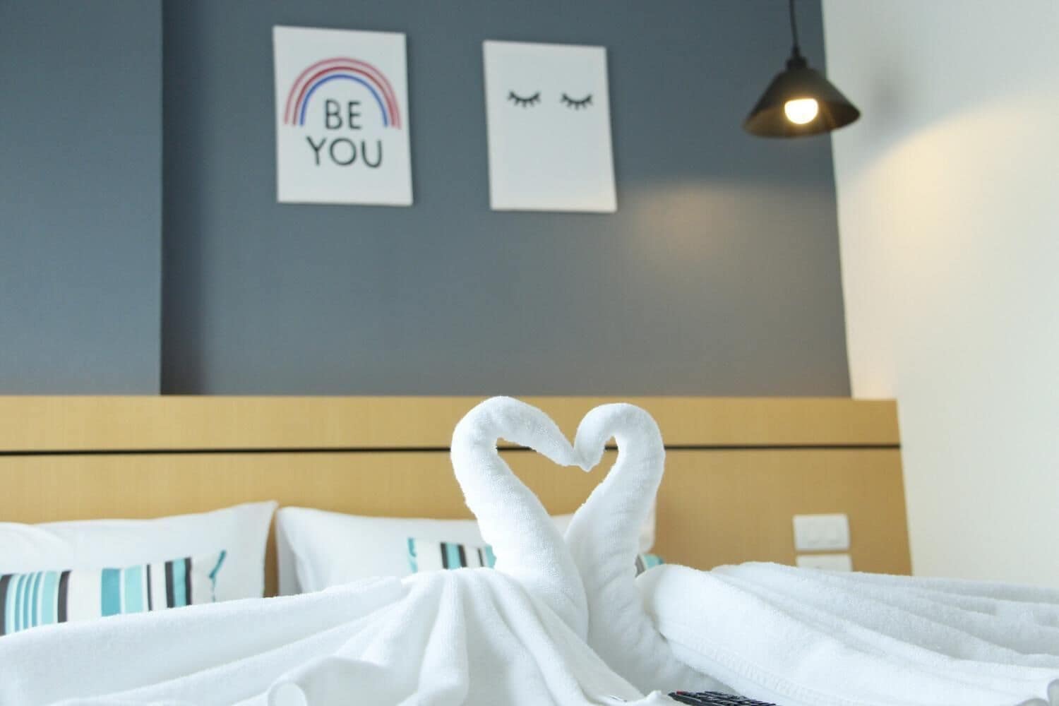 A bed with white towels arranged in a heart shape, a blue wall with two framed pictures, and a hanging lamp.