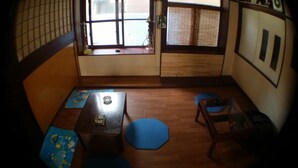 A compact room with a wooden floor, a small table with a blue cushion, a window with a view of the outside, and a door with a traditional design. - Guesthouse Base Okinawa - Hostel (Naha)