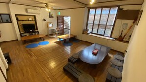 A spacious living area with wooden flooring, a ceiling fan, a large window with a view, and a small kitchen area with a TV and shelves. - Guesthouse Base Okinawa - Hostel (Naha)