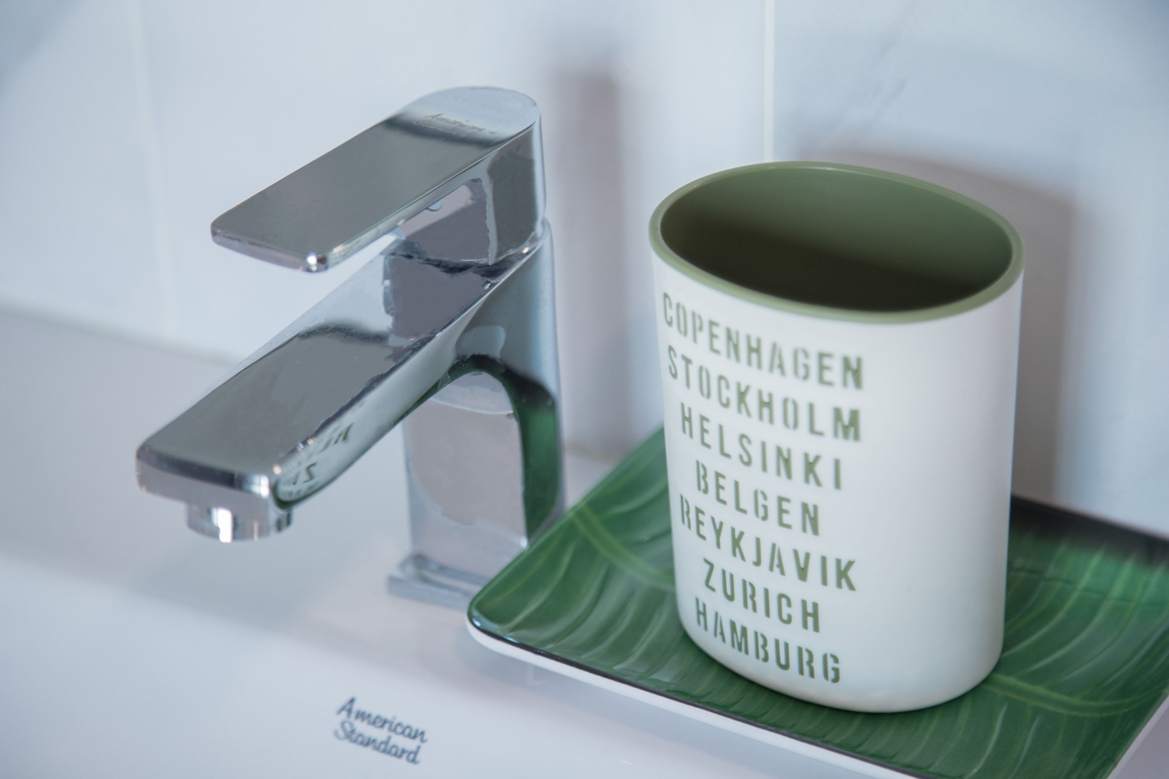A bathroom sink with a modern faucet and a mug with city names on it.