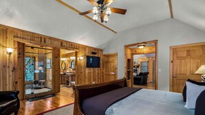 A spacious bedroom with a large bed, wooden walls, a ceiling fan, and a TV mounted on the wall.