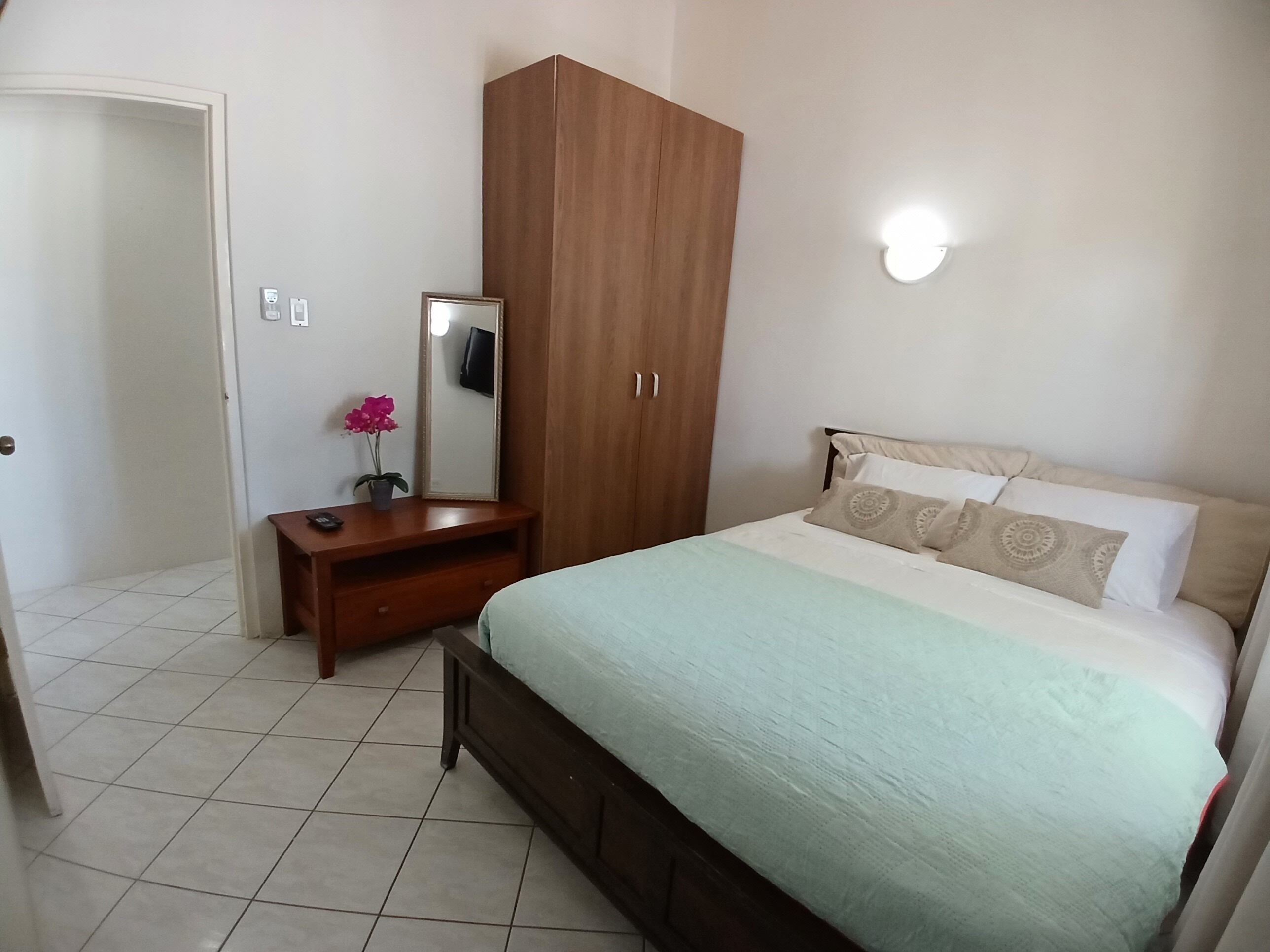 Een slaapkamer met een bed, een houten kledingkast, een nachtkastje met spiegel en een potplant.
