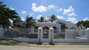 A white building with a gated entrance, surrounded by palm trees and a clear blue sky. - Palm Beach Vacation Villa (Noord)