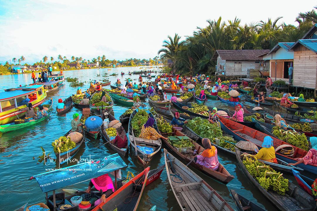Un marché flottant animé, où de nombreux bateaux, chargés de produits frais, sont entourés de maisons traditionnelles et de palmiers.