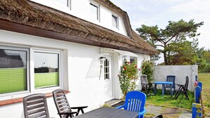 Une maison blanche avec un toit de chaume, une terrasse avec une table et des chaises, et un petit jardin.