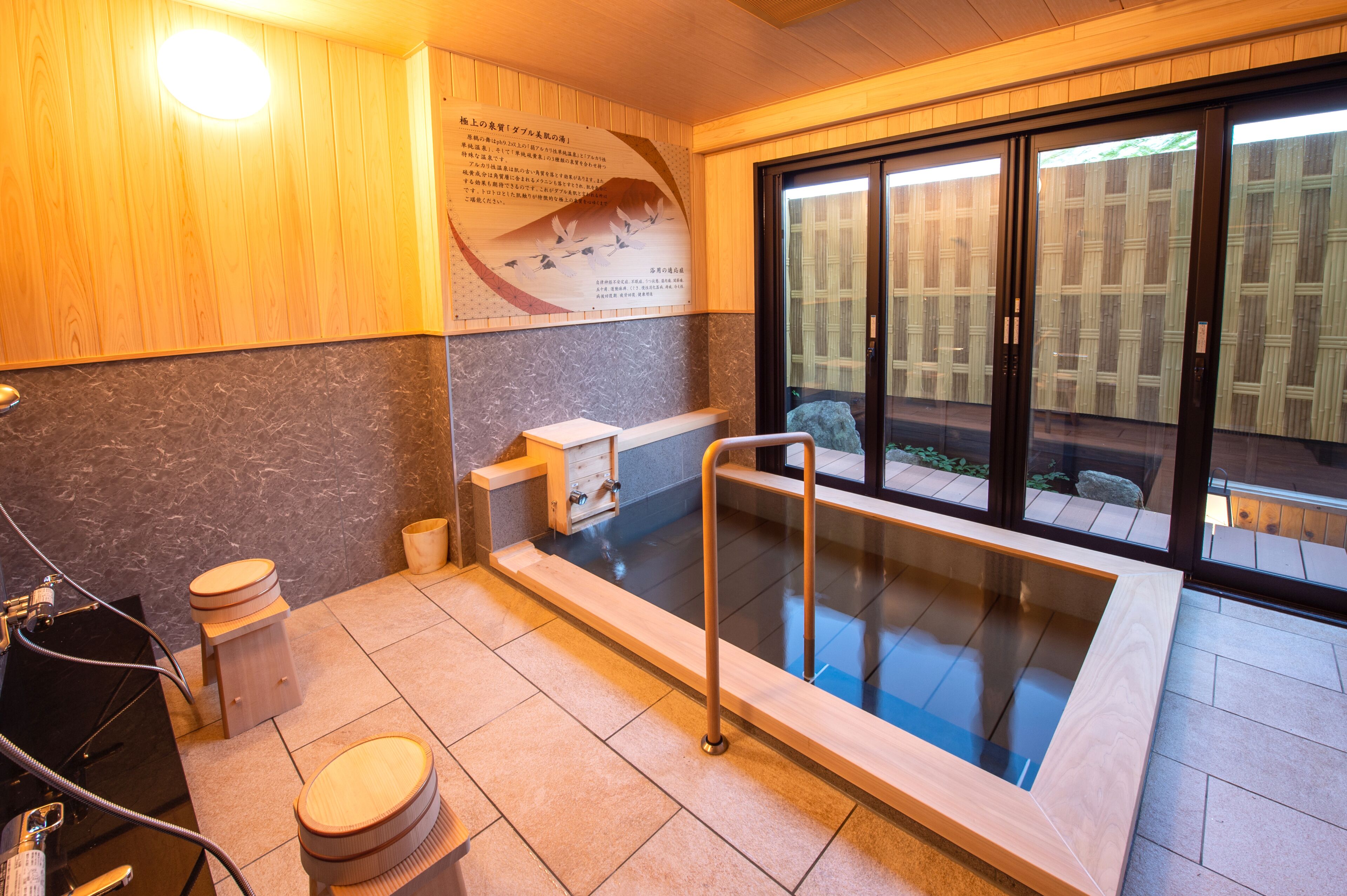 A modern bathroom with a wooden wall, a marble countertop, a glass-enclosed bathtub, and a view of a garden through glass doors.