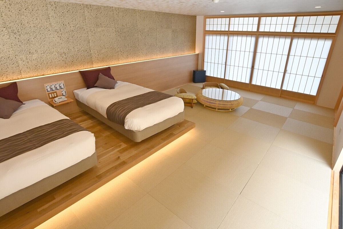 A modern hotel room with two beds, a small round table, and a large window with a view.