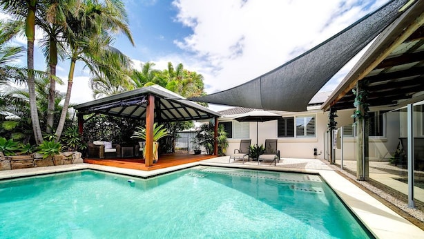 A swimming pool with a covered seating area, surrounded by palm trees and a house with a shaded patio. - Best Location Welcoming Home (Buddina)