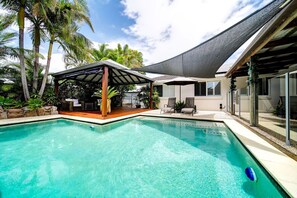 A swimming pool with a covered seating area, surrounded by palm trees and a house with a shaded patio.
