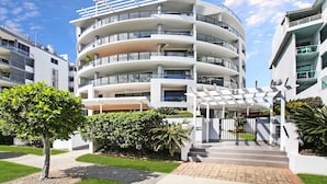 Un edificio residencial de varios pisos con balcones, un acceso cubierto y un jardín bien cuidado.