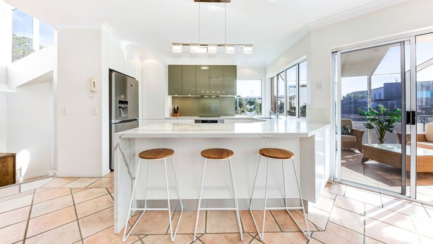 Una cocina moderna con una isla central, taburetes de bar y un amplio ventanal corredero que da a un espacio exterior con mobiliario.