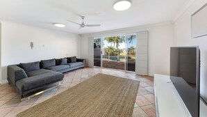 Una sala moderna con un sofá seccional gris, un televisor de pantalla plana, un ventilador de techo y un gran ventanal corredero que da a una zona de piscina.