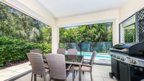 Une terrasse extérieure avec une table et des chaises, un barbecue, et une vue sur une piscine et une végétation luxuriante.