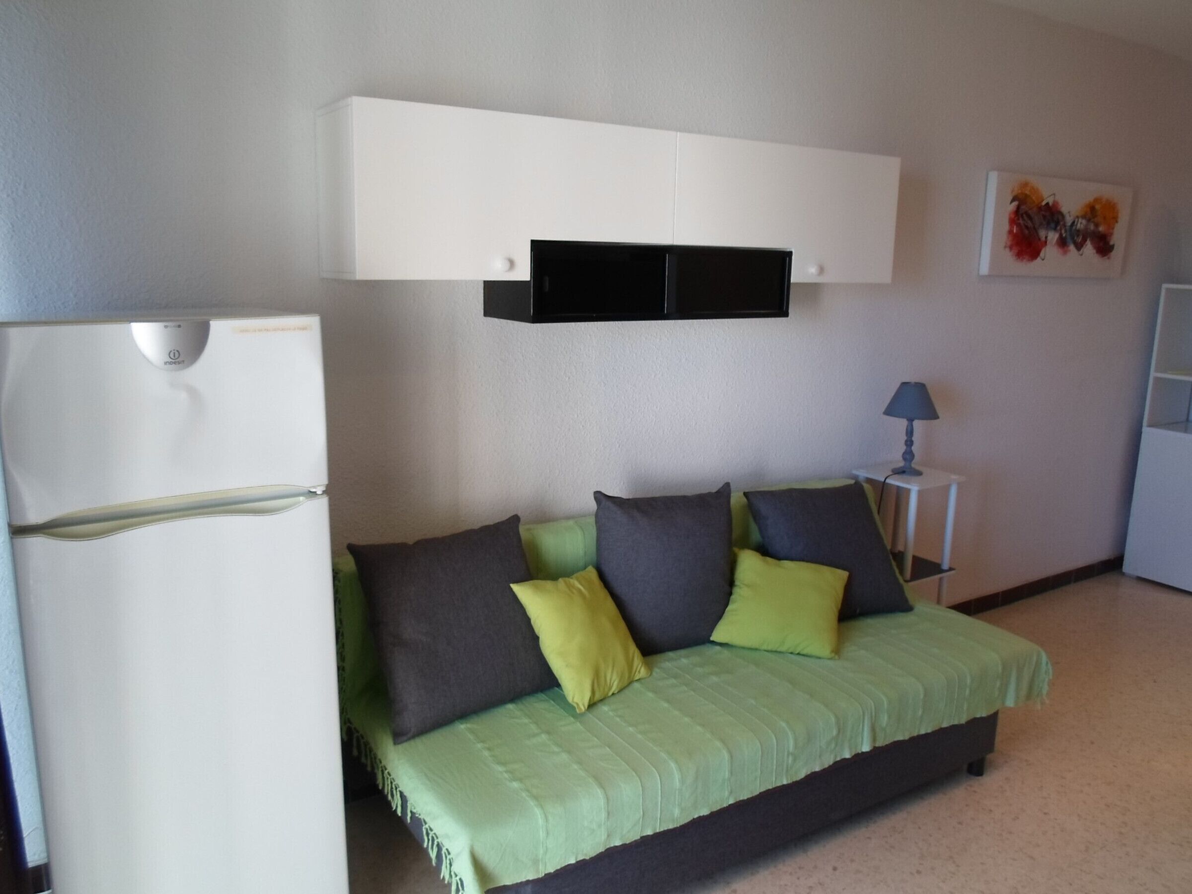 A compact living space with a bed, a white refrigerator, a small table with a lamp, and a wall-mounted cabinet.
