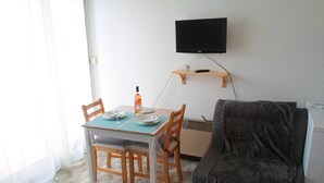 A small, well-lit room with a wooden dining table set for two, a single chair, a sofa, a television mounted on the wall, and a small shelf with a remote control. - Studio near Balaruc Thermal Baths, Air-Con, Pets Allowed (Balaruc-les-Bains)