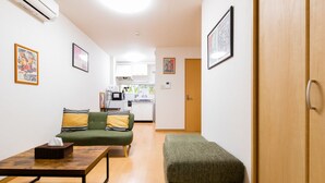 Un espace de vie compact comprenant un canapé, un pouf et une petite table. On aperçoit un coin cuisine au fond et une porte qui donne sur une autre pièce.