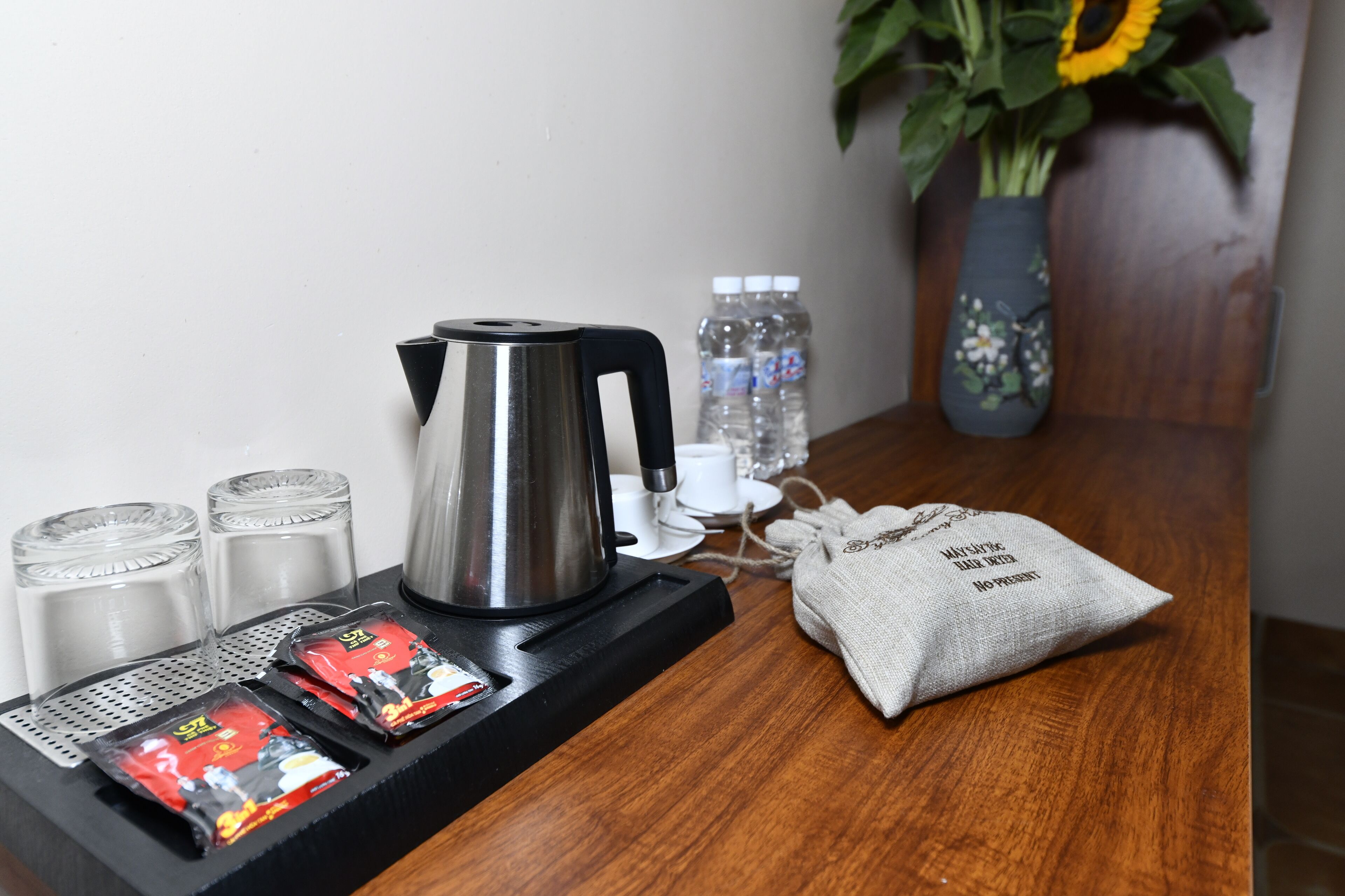 A coffee station with a kettle, cups, and packets of coffee. There are also water bottles and a vase with flowers on the table.