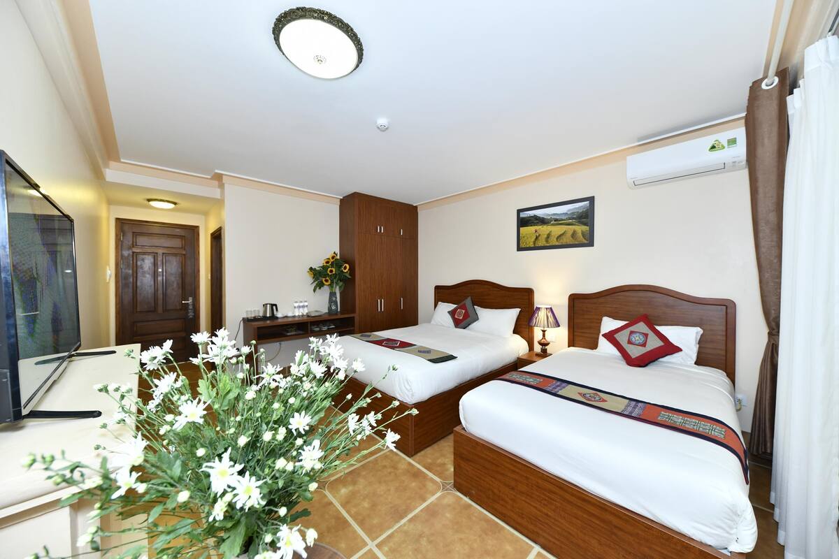 Habitación de hotel con dos camas, televisión, escritorio, un arreglo floral y un cuadro en la pared.