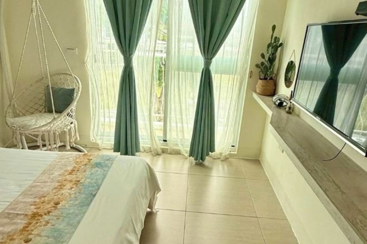 Un dormitorio con cama, una silla colgante, un televisor y un ventanal con cortinas.