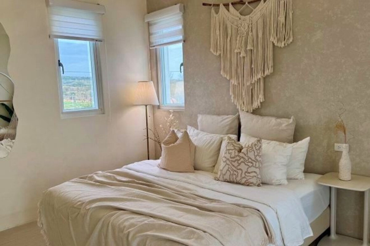 Un dormitorio con una cama, dos ventanas, una mesita de noche y un colgador de macramé en la pared.