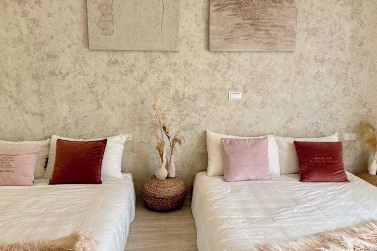 Un dormitorio con dos camas, cada una con ropa blanca de cama y varios cojines en tonos rojo y rosa. Hay una cesta tejida en el suelo entre las camas, y las paredes tienen una textura visible.