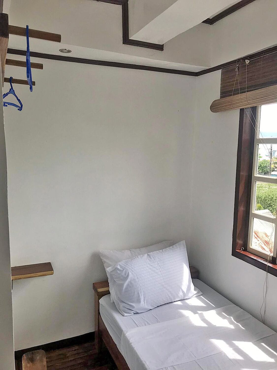 Habitació petita amb un llit individual, panell de fusta al capçal, finestra amb persianes i una prestatgeria fixada a la paret.