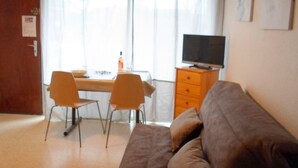 A room with a brown sofa, two chairs, a small table, a television on a wooden cabinet, and a window with curtains.
