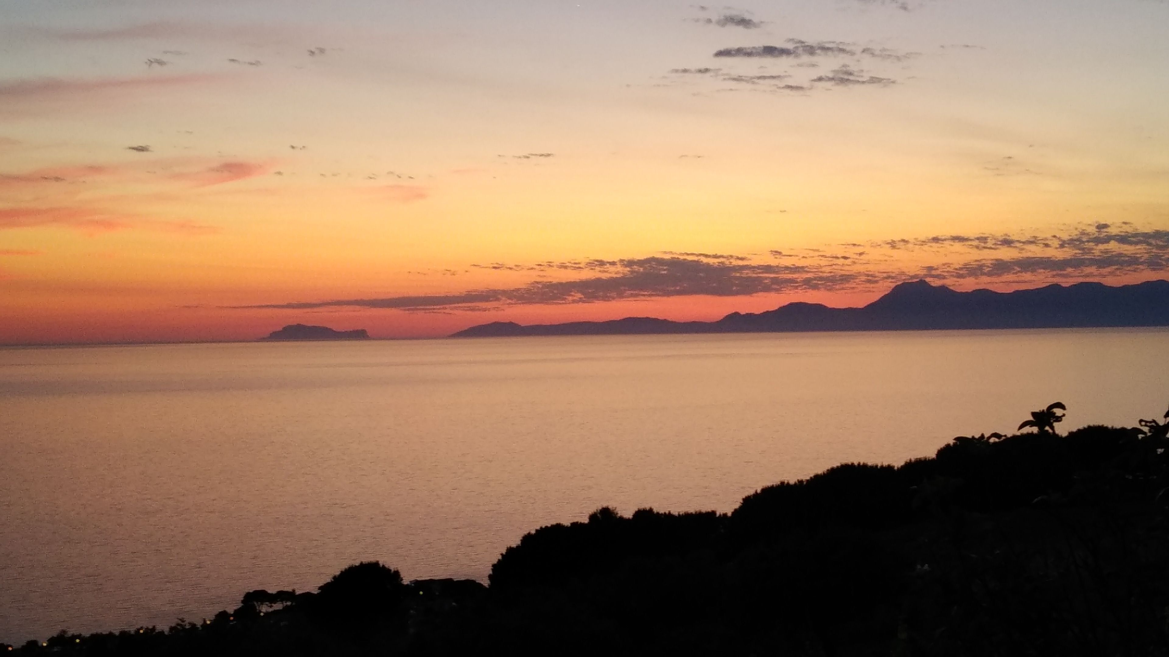 A coastal landscape with a calm sea, distant mountains, and a clear sky during sunset.