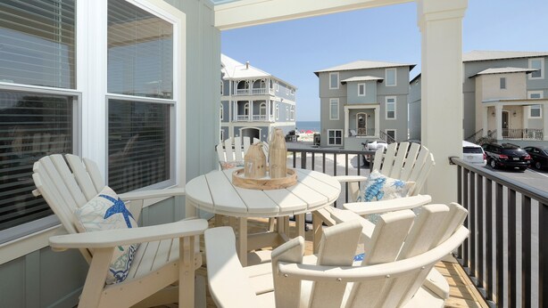 A balcony with white Adirondack chairs, a small round table, and a view of the ocean and buildings in the background. - Beachside Cottage 2BR-3BA. Now booking for Summer! (Orange Beach)